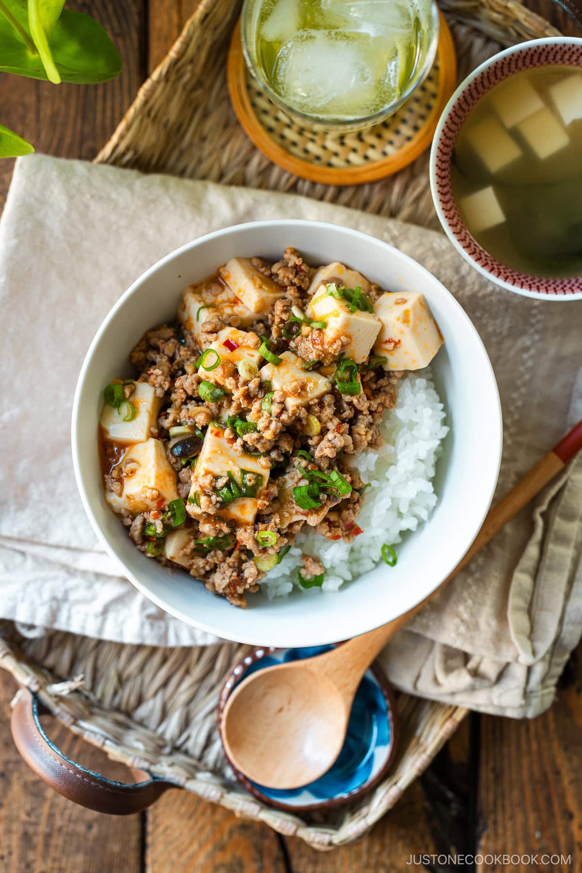 A donburi bowl containing mapo tofu over bed of steamed rice, served with a small bowl of miso soup and a glass of green tea.