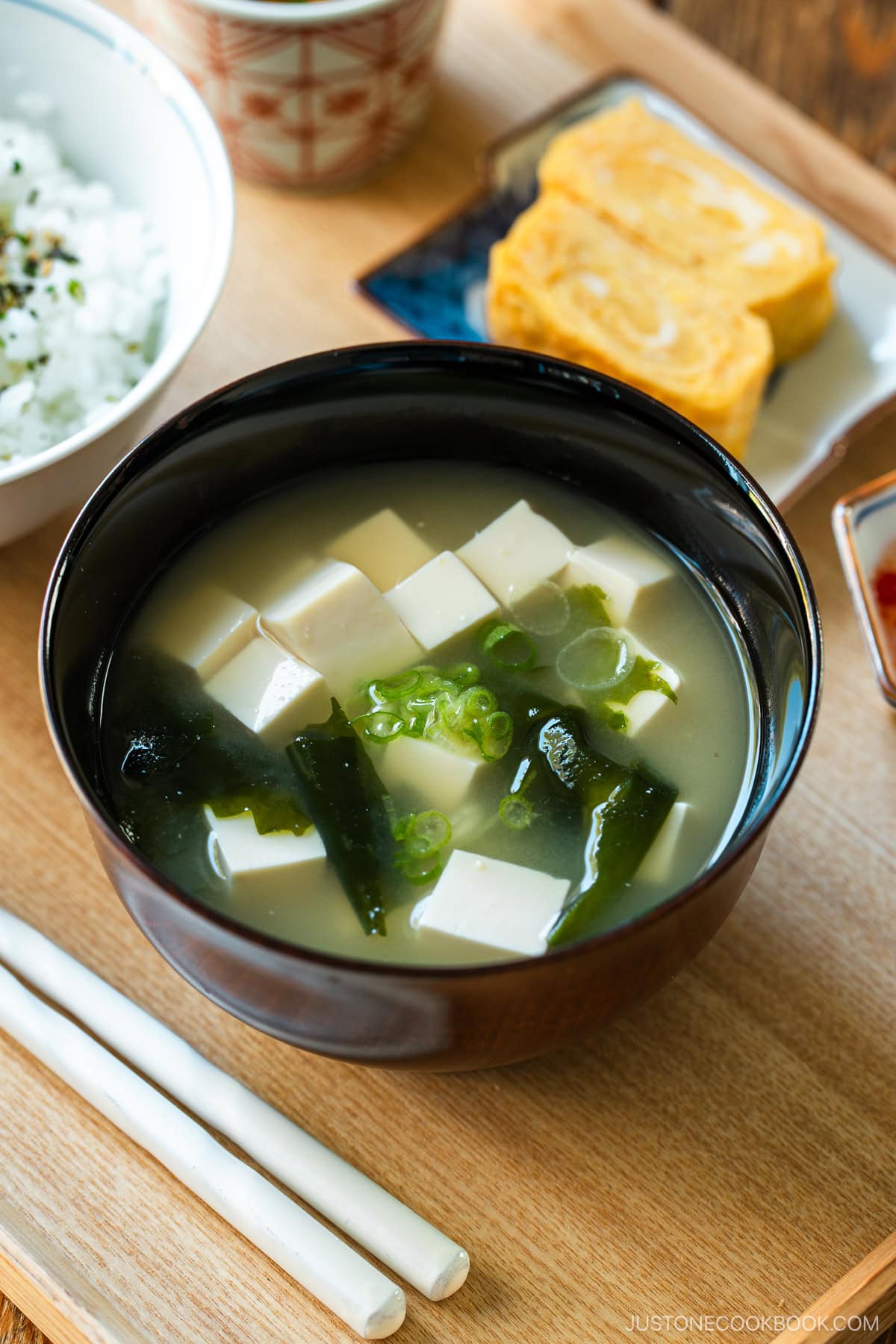 A light-colored wooden tray containing a bowl of miso soup with soft tofu, wakame seaweed, and thinly sliced green onions, along with a bowl of steamed rice, a small plate of tamagoyaki, and a smalll side dish.