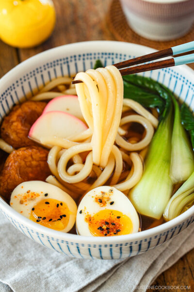 A large blue and white bowl containing udon noodle soup topped with fish cake, bok choy, fish cakes, and jammy soft boiled eggs in a savory dashi broth.