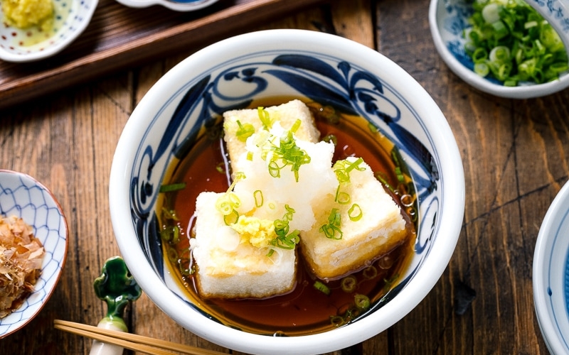 A ceramic bowl containing Agedashi Tofu (Agedashi Dofu) topped with grated daikon, chopped scallions, and grated ginger.