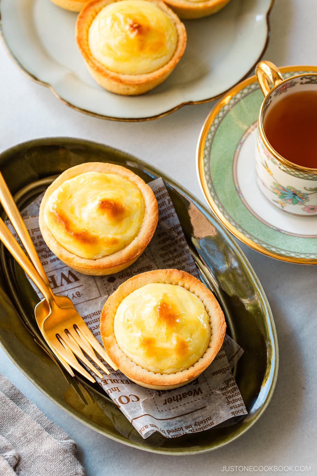 An olive oval plate and a white flower-shaped plate containing baked cheese tarts, served with a cup of tea.