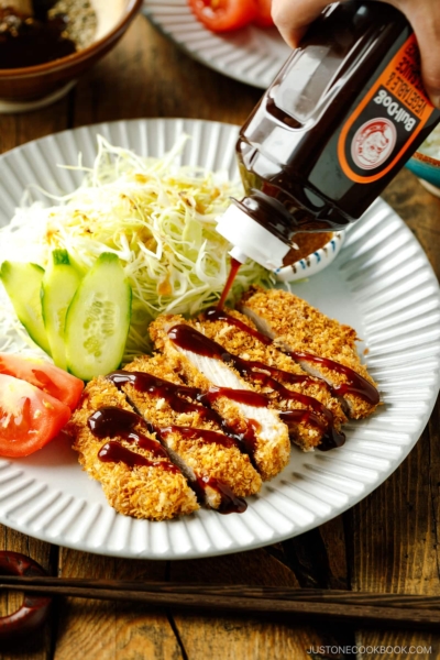 A fluted plate containing baked Tonkatsu drizzled with tonkatsu sauce, shredded cabbage, cucumber slices, and tomato wedges.