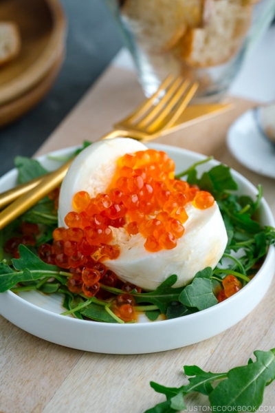 A white plate containing arugula and burrata cheese topped with ikura and served with baguette and yuzu kosho pepper.