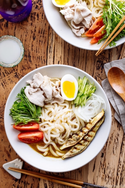 A white bowl containing cold curry udon with sliced pork, boiled egg, and fresh vegetables.