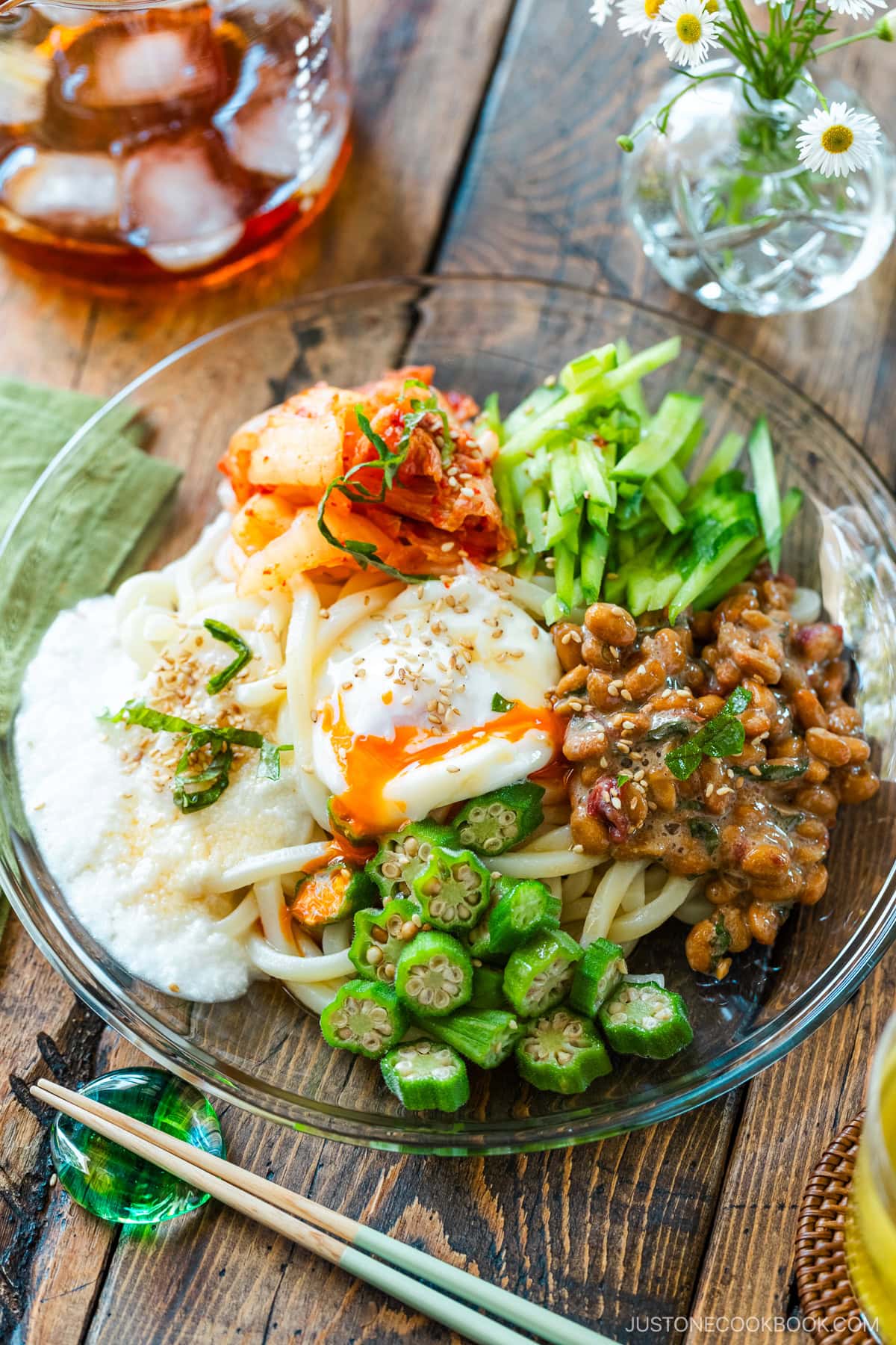 A glass plate containing cold natto udon topped with kimchi, onsen tamago, blanched okra, fresh cucumber, and grated nagaimo.