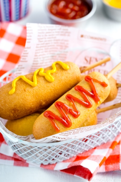 A basket containing corn dogs served with ketchup and mustard drizzle.