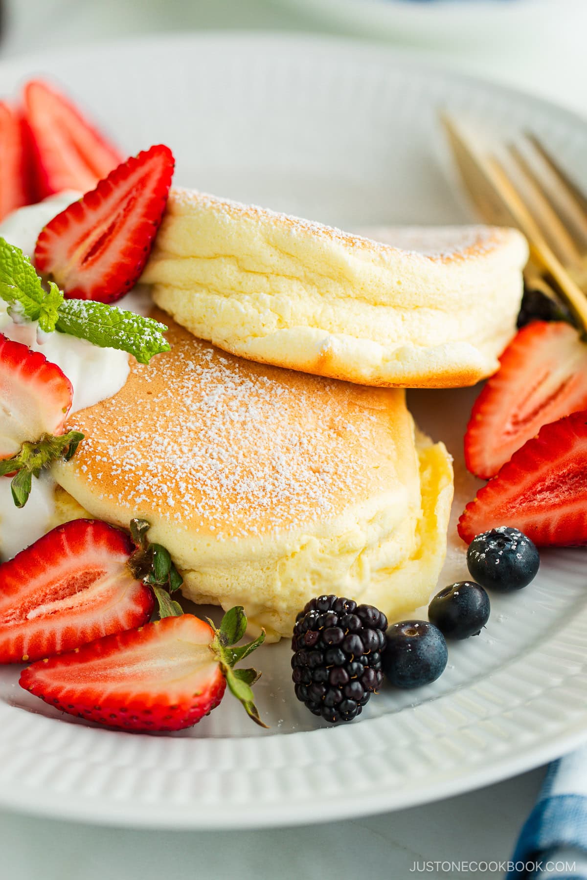 A white plate containing Japanese souffle pancakes dusted with powdered sugar, fresh mixed berries, and whipped cream.