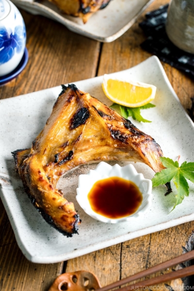 A white Japanese ceramic plate containing Hamachi Kama (Grilled Yellowtail Collar) along with a wedge of lemon and yuzu-soy dipping sauce.