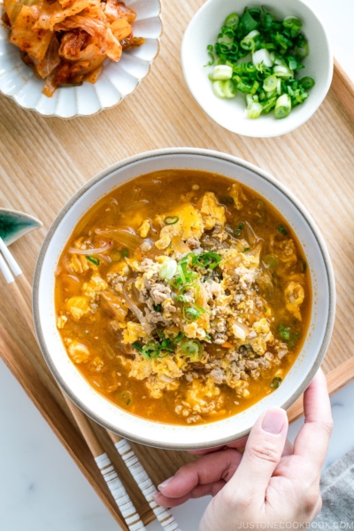 A white bowl containing kimchi soup that's made of Malony glass noodles, kimchi, egg, and pork.