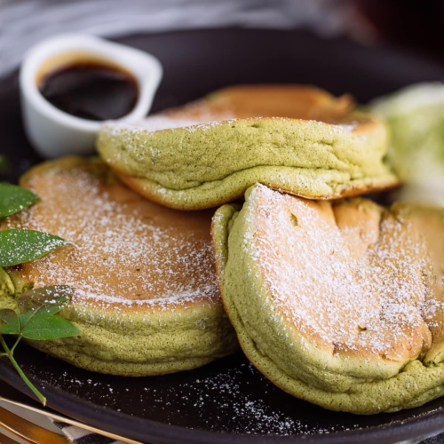 A black plate containing 3 matcha souffle pancakes with fresh whipped cream and maple syrup on the side.