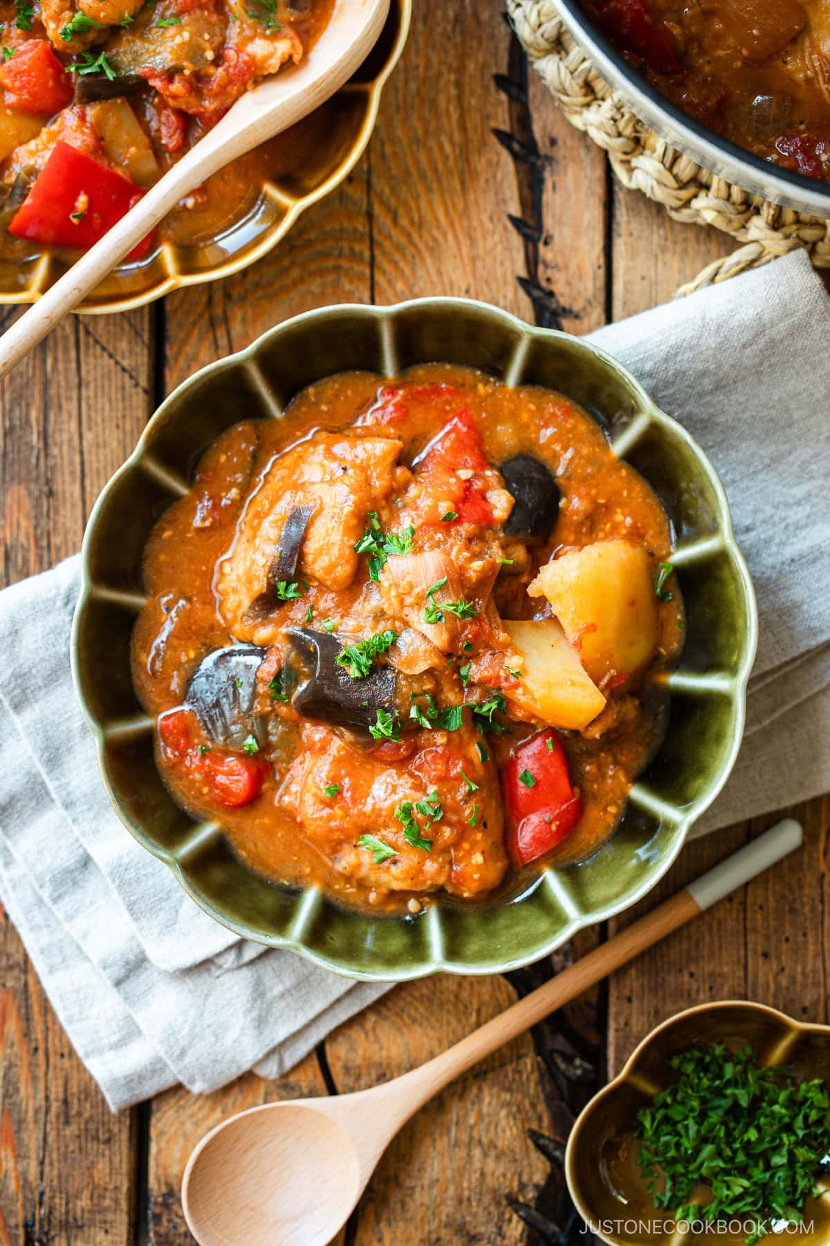 Fluted bowls containing Miso Tomato Chicken Stew garnished with chopped parsley on top, served with a glass of tea, a small dish of parsley, and a pot of the stew on the wooden table.