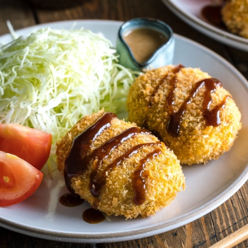 A white plate containing Japanese Croquette (Korokke) served with Tonkatsu sauce and shredded cabbage on the side.