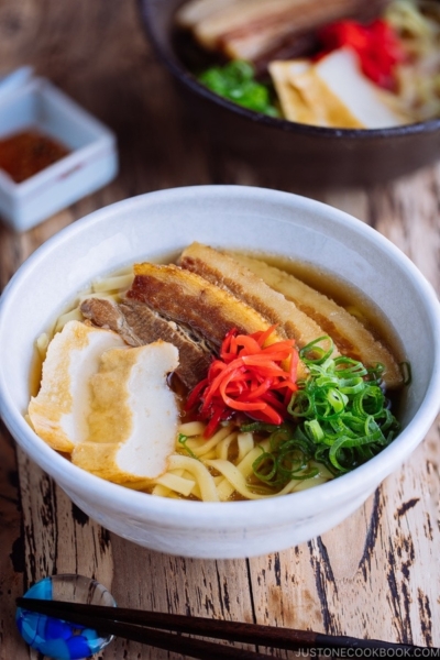 A white bowl containing Okinawa Soba and delicious Japanese dashi and pork broth, topped braised pork belly, fish cakes, green onion, and red pickled ginger.