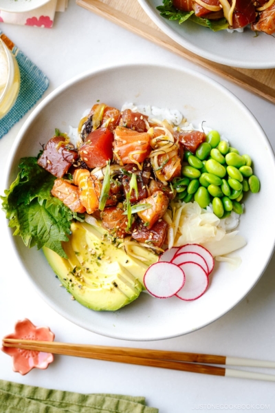 A bowl containing ahi tuna and salmon poke along with edamame, avocado, sushi ginger, and radish over bed of steamed rice.