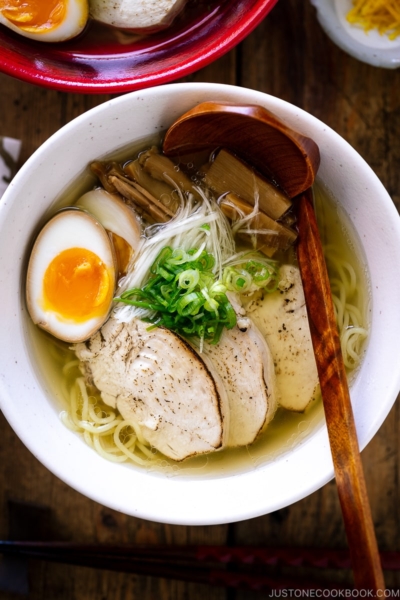 A white bowl containing Shio Ramen, topped with Chicken Chashu, Ramen Egg, bamboo shoot, white negi, and green onion.