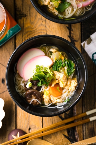 A black bowl containing somen noodle soup with vegetables, shiitake mushrooms, egg, kamaboko fish cake, garnish with green onion.