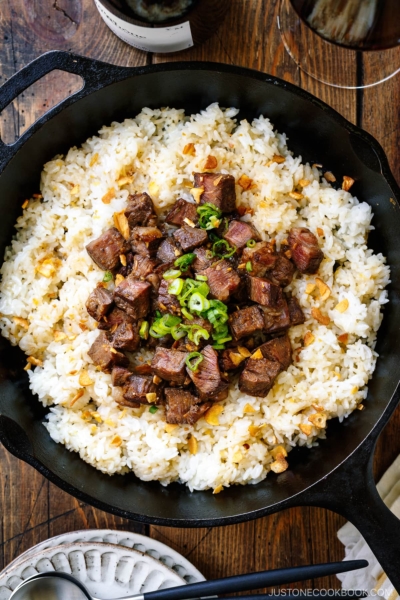 A cast-iron pan containing Steak Garlic Rice topped with chopped scallions and garlic chips.