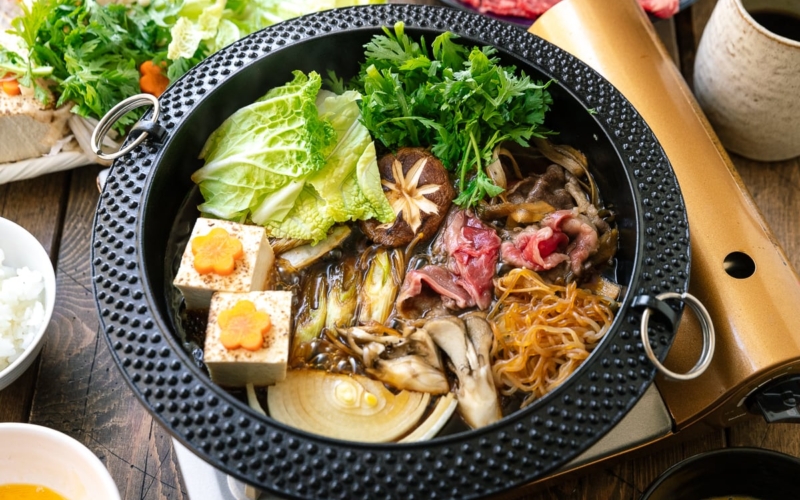 Japanese round cast iron pan containing Sukiyaki, a Japanese hot pot dish where marbled beef, tofu, and vegetables are simmered in sweetened soy sauce broth.