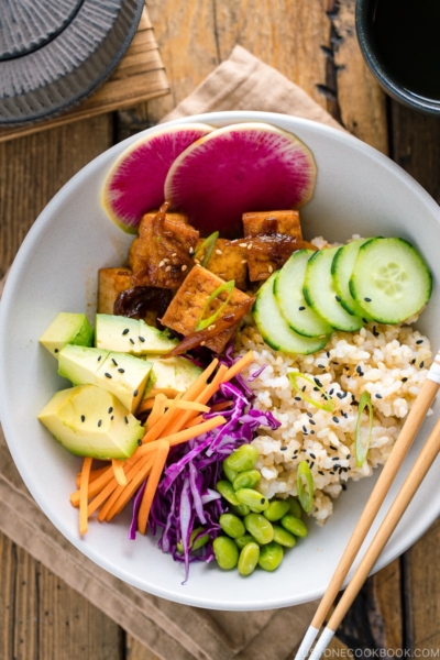 A white bowls containing savory pan-fried tofu, cucumber, avocado, edamame, carrot, red cabbage, and watermelon radish over brown rice.