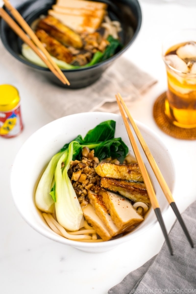 A white bowl containing Vegetarian Udon Noodle Soup topped with bok choy, fried tofu, minced mushroom, and crispy eggplant.