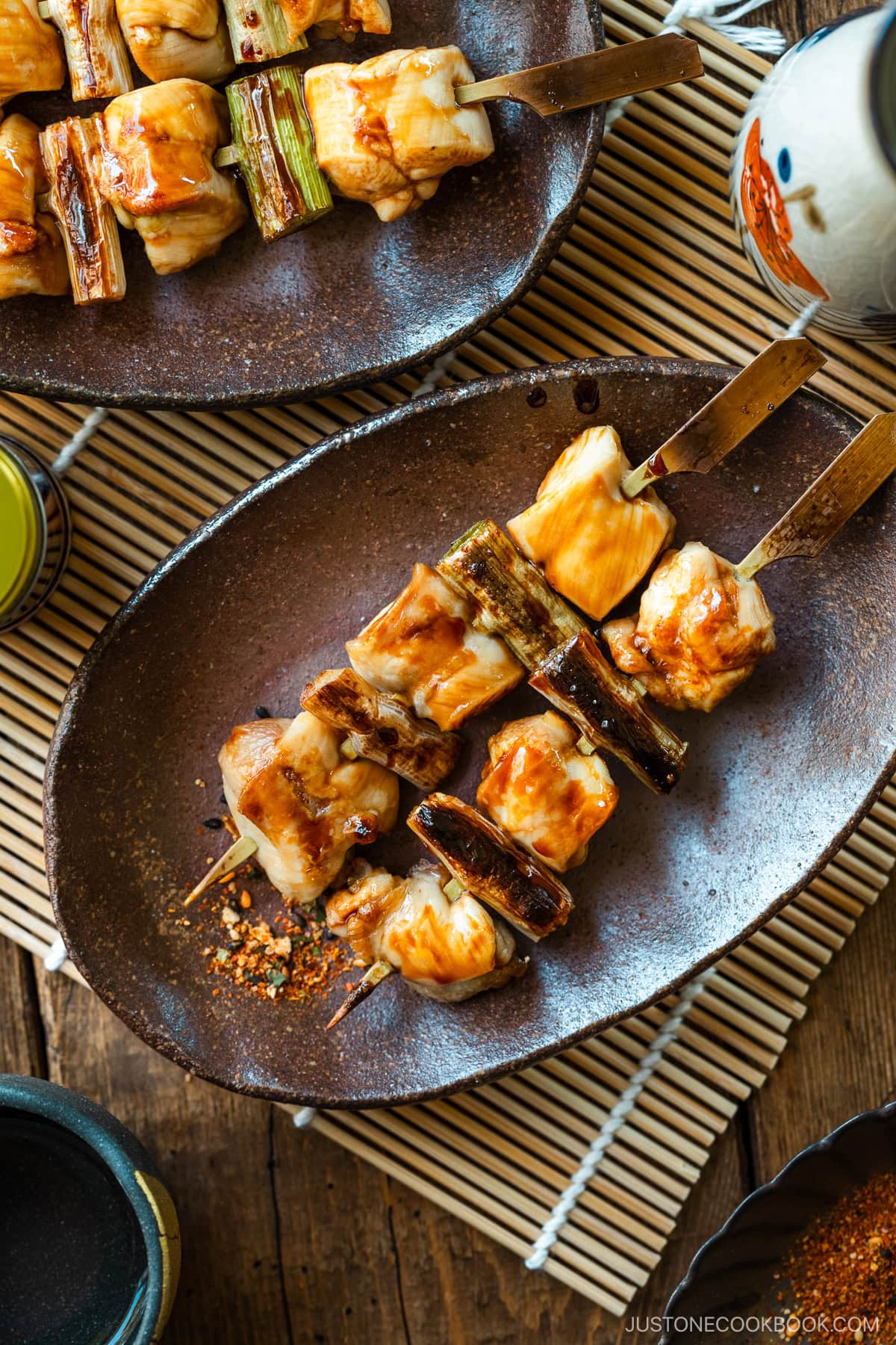 Oval rustic plates containing yakitori, Japanese grilled chicken and scallion skewers, coated with savory yakitori sauce and served with shichimi togarashi on the side.