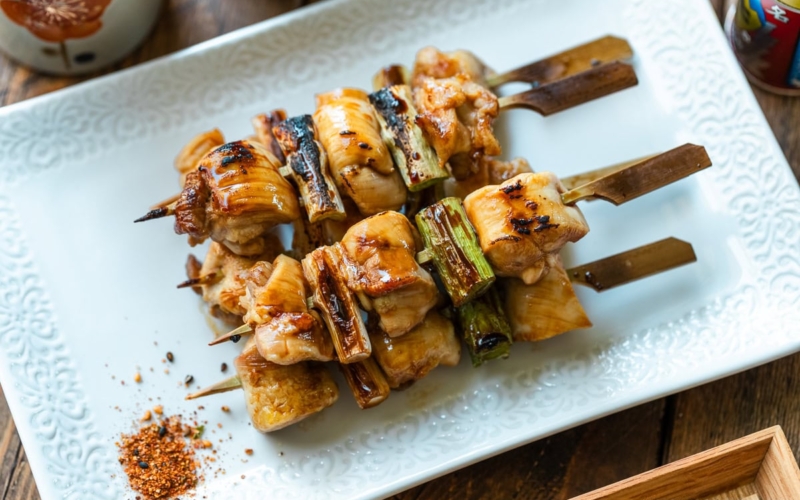 A white rectangular plate containing yakitori, Japanese grilled chicken and scallion skewers, coated with savory yakitori sauce and served with shichimi togarashi on the side.